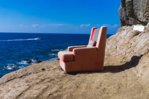 in a show of utter randomness there is an armchair sitting on a rocky cliff overlooking the ocean. The sky is blue.