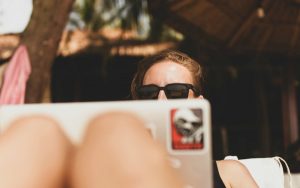 A lady is sitting behind a laptop with her knees up to signify remote work conditions. She has sunglasses on and is feeling casual.
