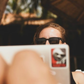A lady is sitting behind a laptop with her knees up to signify remote work conditions. She has sunglasses on and is feeling casual.