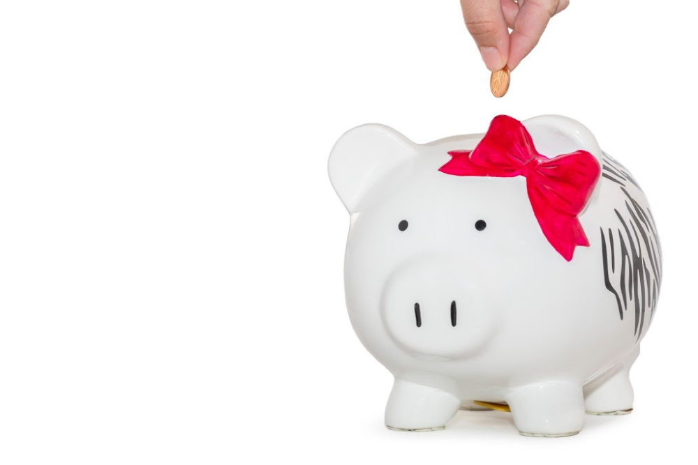 Illustrating the idea of saving on a marketing budget with a white ceramic piggy back wearing a bow. There is a hand dropping coins into the piggy bank.