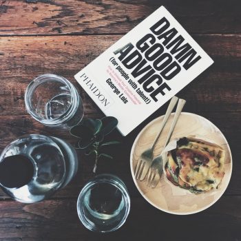 On a wodden table, a flat lay of toast, two glasses of water, a bottle and a book entitled damn good advice lie. It's to denote the whole advice culture with cafe side that business and life coach people are enamoured with.