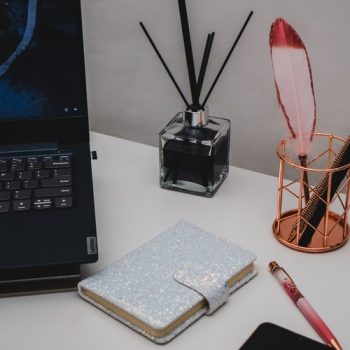 Looking at the desk of a conceptual copywriter there is a laptop, diary with clasp, an oil diffuser and a bottle of pens ready to go.