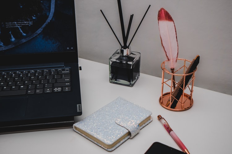 Looking at the desk of a conceptual copywriter there is a laptop, diary with clasp, an oil diffuser and a bottle of pens ready to go.