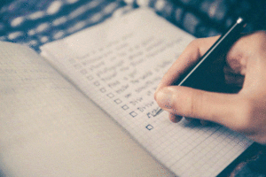 A hand is engaged in some business planning in a journal. The hand is holding a pen. There are faint check box items and a list.