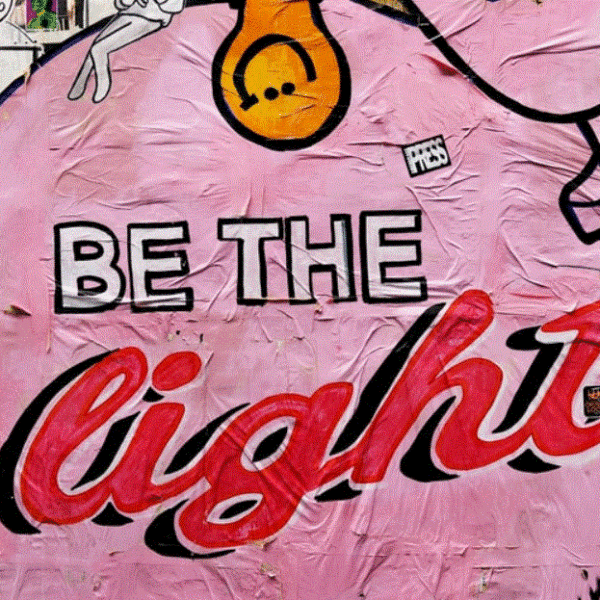 A graffiti pasteup is pink and features retro fonts. It reads be the light just like you can be with digital content marketing.. It has a smiley.